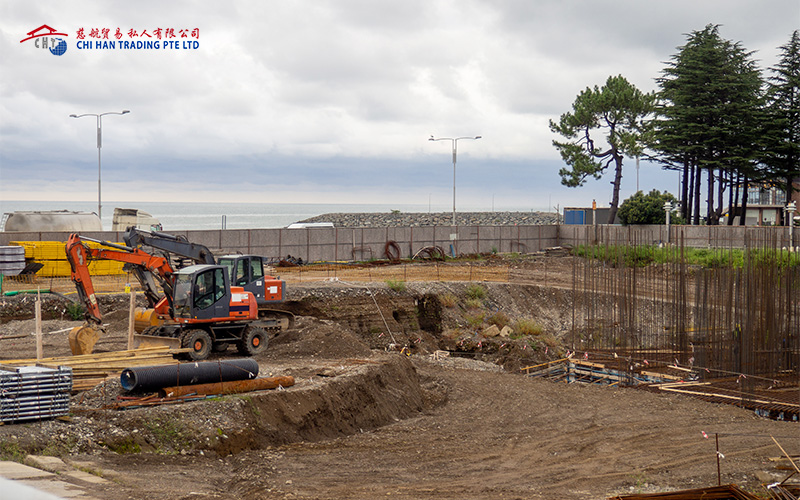 Excavators at a coastal construction site under cloudy skies.